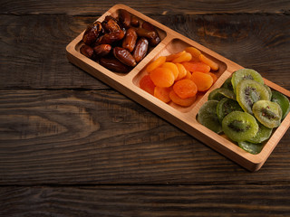 Dates, dried apricots and kiwis in a Compartmental dish on a dark wooden table.