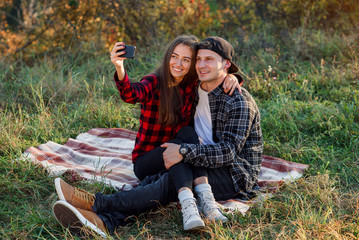 Naklejka na ściany i meble Young happy hipster couple making selfie using smart phone while sitting on the lawn in spring park. Stylish modern couple have a video call with their friends sitting in the nature.