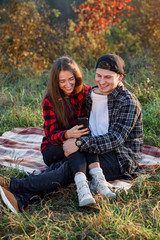 Beautiful girl sits on her boyfriend lap and using smart phone on the lawn in the park. Hipster modern couple have a chatting with their friends sitting in the nature.