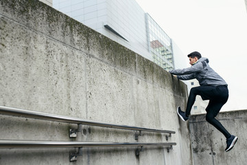 Stylish sportsman use architecture of urban area for his workout. He climbs at concrete wall, in the background modern city buildings.