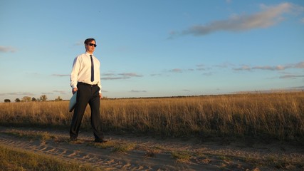 Naklejka na ściany i meble businessman in sunglasses goes down country road with briefcase in his hand. agricultural business concept. The entrepreneur works in a rural area. farmer inspects the land at sunset. Slow motion