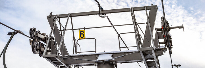 Steel ropes over wheels in mechanism on top of ski chairlift support column, number 6 on yellow plate, bright sky background, wide banner