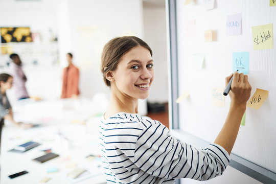 Waist up portrait of contemporary young woman drawing on whiteboard in office and smiling at camera, copy space