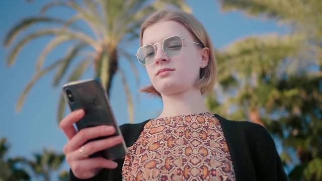 Portrait of young attractive trendy teenager girl in stylish sunglasses waiting for her friends using smartphone to check social networks, mail, Instagram background palms blue sky