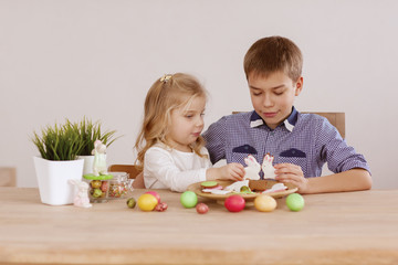 Obraz premium A girl with an older brother are sitting at the holiday table and laying out cookies and Easter eggs