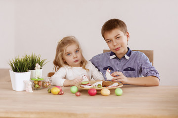Obraz na płótnie Canvas A girl with an older brother are sitting at the holiday table and laying out cookies and Easter eggs