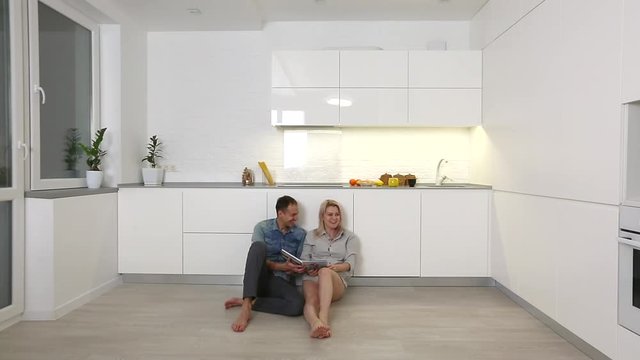 Joyful lovely couple browsing pictures photoalbum, sitting on the kitchen floor, having warm and nice talk