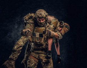 Combat conflict, special mission, retreat. Soldier special forces rescue his wounded teammate carrying him on his shoulders from the battlefield. Studio photo against a dark wall