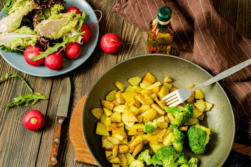 vegetarian meal of fried potatoes and broccoli with salad and fresh radish on a wooden table