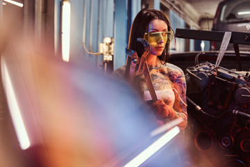 Female model with tattooed body wearing protective goggles posing with a big wrench next to a car engine suspended on a hydraulic hoist in the workshop. Photo with red light illumination