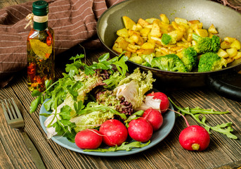 vegetarian meal of fried potatoes and broccoli with salad and fresh radish on a wooden table