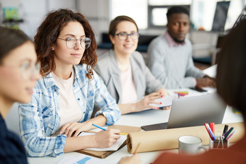 Group of contemporary people working in meeting focus on curly haired young woman in foreground,...