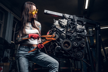 Stylish female model with tattoed body repairs a car engine suspended on a hydraulic hoist in the workshop.