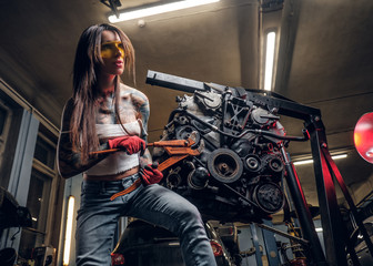 Obraz na płótnie Canvas Stylish female model with tattoed body repairs a car engine suspended on a hydraulic hoist in the workshop.