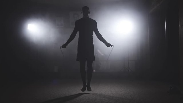Boxer does some jump rope exercises in dark room under light. Fighter training in smoky studio. Silhouette on dark background