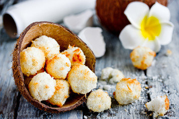 Obraz na płótnie Canvas Homemade coconut candies from coconut chips, round crumbly coconut cookies in half coconut shell on wooden table with plumeria flower