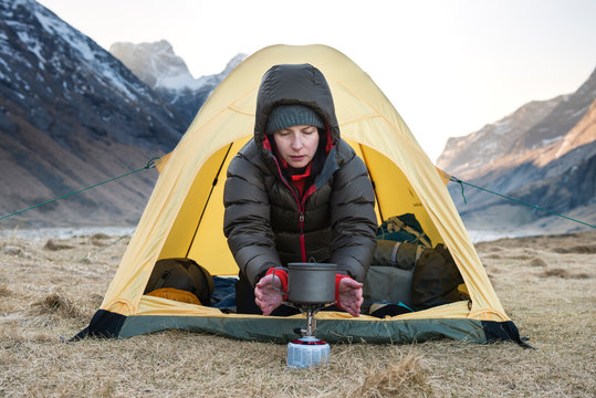 female hiker warms hands with stove on cold morning wild camping at Horseid beach, Moskenes&oslash;y, Lofoten Islands, Norway