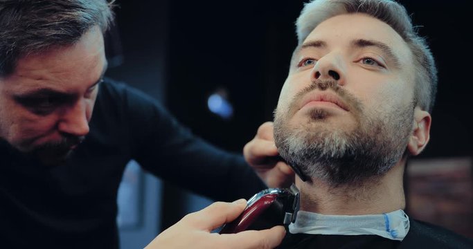 Adult man cuts his beard in the barbershop. Hairdresser dub beard machine for cutting. Portrait view.