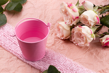 A bouquet of beautiful roses stands in a small bucket on a lace ribbon on a pink craft background with space for text