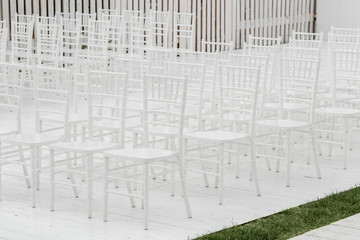 Rows of empty white chairs sitting on a wooden floor. Wedding chairs with flowers at ceremony outdoors