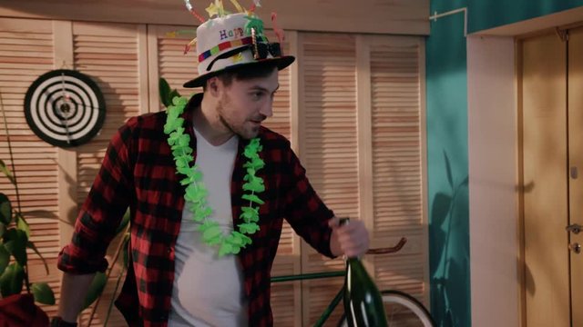 Drunk guy in funny B-day hat and Hawaiian wreath holding bottle of alcohol in hand and drinking. Sloshed young man falling on bed. Drinker coming home