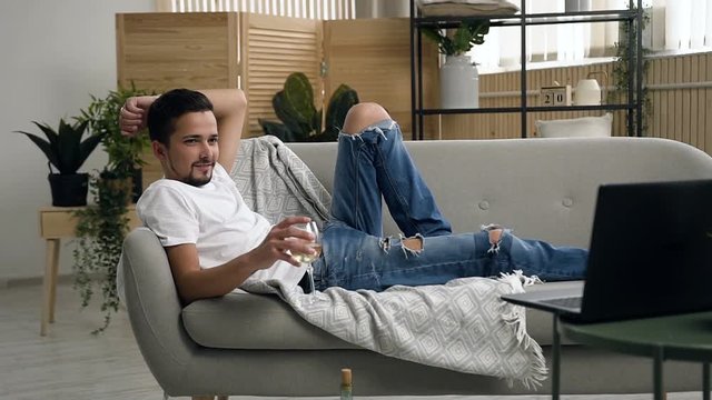 A young attractive man lying the couch relaxing and drinks sparkling white wine behind watching movie in laptop in the living room