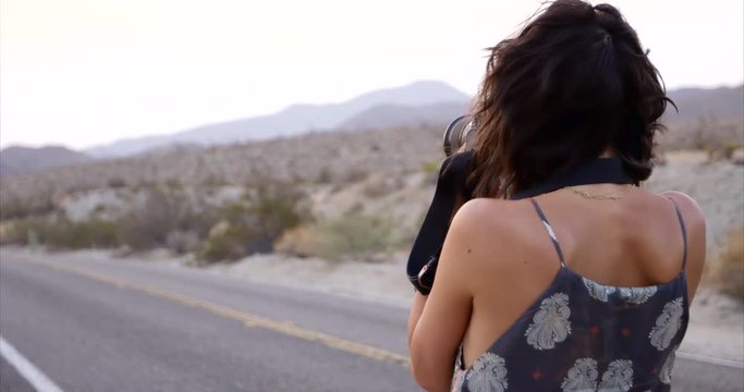 Stylish female photographer taking photos along desert highway - from behind rotating shot