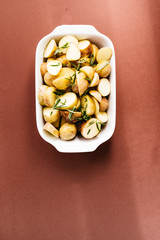 Baby jacket potatoes with herb and garlic oil dressing, on a white ceramic container over a brwon board, top view, baked Potatoes in Pan, Cooking with Garlic & Fresh Rosemary.