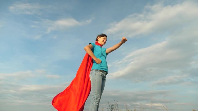 girl dreams of becoming a superhero. young girl standing in a red cloak expression of dreams. beautiful girl superhero standing on the field in red cloak, cloak fluttering in the wind. Slow motion
