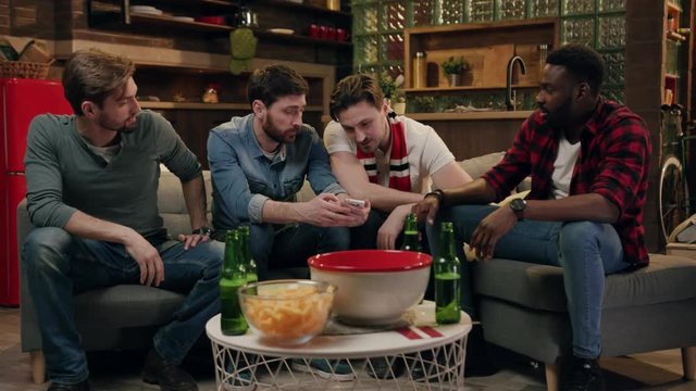 Young multiracial male friends using digital device, discussing the image on a screen. Friendship concept, home party, beer beverages, snacks. Modern technology concept, social networks, surfing