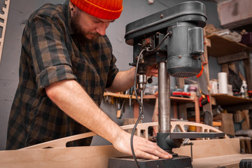 Close up of experienced carpenter in work clothes and small buiness owner  is carving a wooden board on an  modern  hand drill in a light workshop side view, in the background a lot of tools