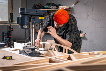 A man carpenter in a hat and a shirt  is carving a wooden board on a large drilling machine in a...