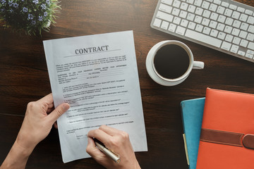 Fototapeta na wymiar contract and agreement concept, man holding pen to sign contract application paper with coffee cup keyboard and notebook on wood desk in office bank.
