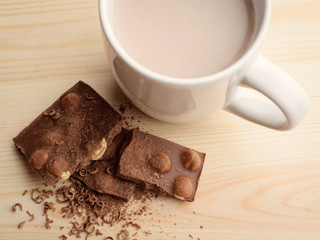 Hot cocoa in white cup stands on wooden table near some pieces of chocolate bar with hazelnuts seasoned with cocoa powder and grind chocolate
