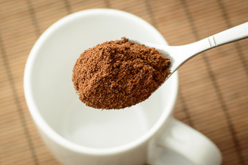 Spoon with ground coffee over the white empty cup against a brown bamboo background. Making coffee