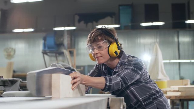 Medium shot of female joiner wearing safety headphones and goggles cutting piece of wood with electric jointer