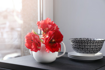 Teapot with beautiful flowers and dinnerware on table near window