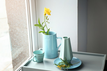 Jug with beautiful flower and tableware on tray near window