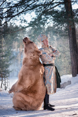 A man in traditional national costume of a Russian prince stands by a brown bear in a winter...