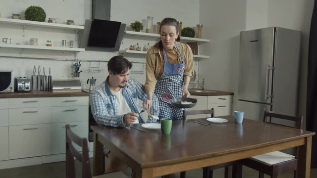 Disgruntled man sitting at kitchen table attacking wife with complaints about prepared breakfast. Irritated husband with disgust looking at scrambled eggs, swearing, offending woman with his behavior.