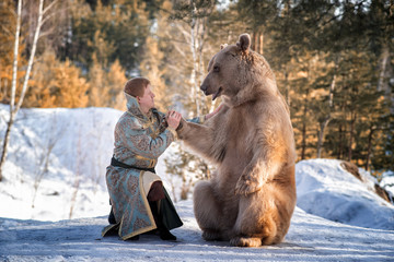 A man in traditional national costume of a Russian prince sits on one knee and holds a brown bear by the paw in a winter forest.