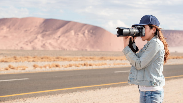 Young Asian woman traveler and photographer holding camera taking photo of sand dune near the road in Namib desert of Namibia, Africa. Travel photography concept