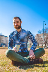 The young guy with beard and in shirt sits on grass having crossed legs in the spring park and drinks coffee from paper glass. Mood.