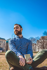 Fototapeta premium The young guy with beard and in shirt sits on grass having crossed legs in the spring park and drinks coffee from paper glass. Mood.