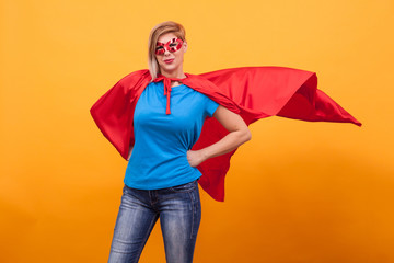 Young woman in superheros costume standing proudly with her red cape in the air over yellow...