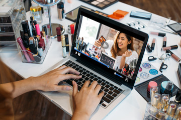 Crop hands of woman using laptop and watching beauty and makeup video online with makeup kits at table - Asian woman blogger