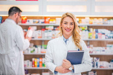 Portrait of a smiling blonde female healthcare worker in a pharmaceutical store, colleague working...