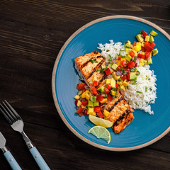 Grilled salmon fillet with fruit sauce made of mango, bell pepper, onion and avocado with basmati rice garnish on a blue plate. Delicious seafood meal. Top view shot above.