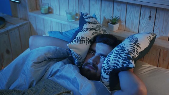 Disgruntled young bearded man suffering from sleeplessness. Guy looking up, rolling eyes, covering ears with pillows. Indoors. Insomnia concept, bedroom, bedtime, disturbed,  exhausted, frustrated