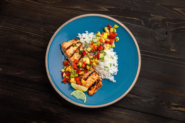Delicious grilled salmon steak on a blue plate with jasmine rice and sauce made of mango, bell pepper, onion and avocado. Top notch food. Top view shot above.